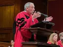 Former Harvard President Lawrence S. Bacow in a red academic robe gestures passionately at a podium, with seated audience members behind.