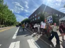 Protesters holding signs march along a sunny street in front of a building.
