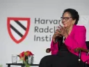 A woman in a pink blazer smiles while seated on stage at a Harvard event.