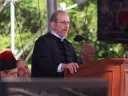 Harvard President Alan Garber in academic robes speaks at a podium outdoors, with a blurred audience behind him.