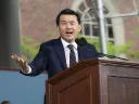 Ronny Chieng wearin a suit speaks passionately at a wooden podium outdoors.