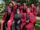 Group of seven individuals in academic regalia, smiling outside on a sunny day.