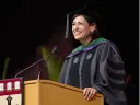 A smiling woman in academic regalia speaks at a podium during a graduation ceremony.