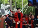 A speaker in academic regalia stands before an audience during a ceremony outdoors.