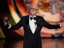 A man in a tuxedo gestures while holding an award on a stage.