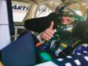 Racing driver gives a thumbs up from inside a car, wearing a helmet and safety gear.