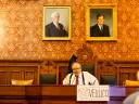 A man sitting at a wooden table in a formal setting with portraits on the wall.