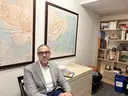Man in a gray blazer sitting in an office with world maps on the wall and a bookshelf behind.