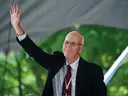 A man in a suit smiles and waves while standing outdoors under a tent.