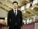 A man in a suit stands confidently on an ice rink, smiling, with a sports arena in the background.