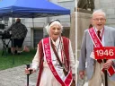 Older woman in a white outfit with a cane and red sash, alongside an elderly man in a gray suit holding a sign.