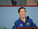 Smiling astronaut in blue jumpsuit speaking at a podium, colorful background.