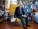 Harvey Mansfield seated in a bright yellow chair, surrounded by bookshelves and cozy decor.