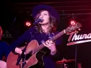 Singer performing on stage with a guitar, wearing a hat, and surrounded by band instruments.