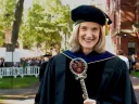 Katie O’Dair in academic regalia holds a ceremonial staff outdoors at a graduation ceremony.