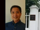 A man smiling in a blue shirt next to a white gate with a plaque and greenery.