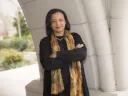 A woman with a black sweater and patterned scarf stands under an archway.