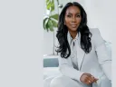 A woman with long, wavy hair wearing a white suit, seated in a bright, modern room.
