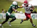 A football player in a white jersey evades a defender in a green uniform on the field.
