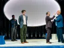 Three actors perform on stage, with one actor interacting closely with another, set against abstract backgrounds.