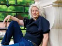 A woman with curly blonde hair sits casually against a stone wall, wearing a black top and jeans.