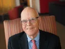 A smiling elderly man in glasses sits comfortably in a striped armchair.