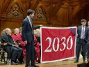 Two men hold up a red banner with "2030" written on it in white in a wood paneled room