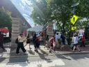 A group of protesters holding signs marches past a gated entrance on a sunny day.