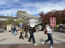 Protesters march holding signs in a public square with buildings and trees in the background.