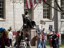 Crowd protesting near a statue, holding signs with various messages; flags above.