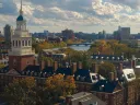 Panoramic view of Harvard University with autumn foliage and a river under a cloudy sky.