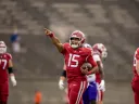A football player in a red uniform points while holding a ball, with teammates nearby.