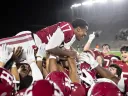 Harvard team, wearing red uniforms, cheer and celebrate together by raising player #17 into the air