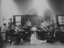 Several Harvard Computers and Radcliffe students pose for a photograph together, seated at desks and sitting on the floor