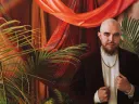 A formal portrait or José Olivarez in a gold chain and velvet suit jacket, standing amid red drapes and palm plants.
