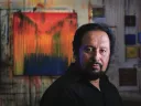 Painter George Oommen in his studio. His painting <em>Visions of Kerala 2</em> (acrylic on  canvas, 2005) hangs behind him. 