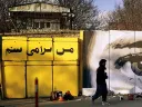 Along a city street, mural on a concrete wall shows two men shaking hands, with a large pair of watchful eyes in  the background.